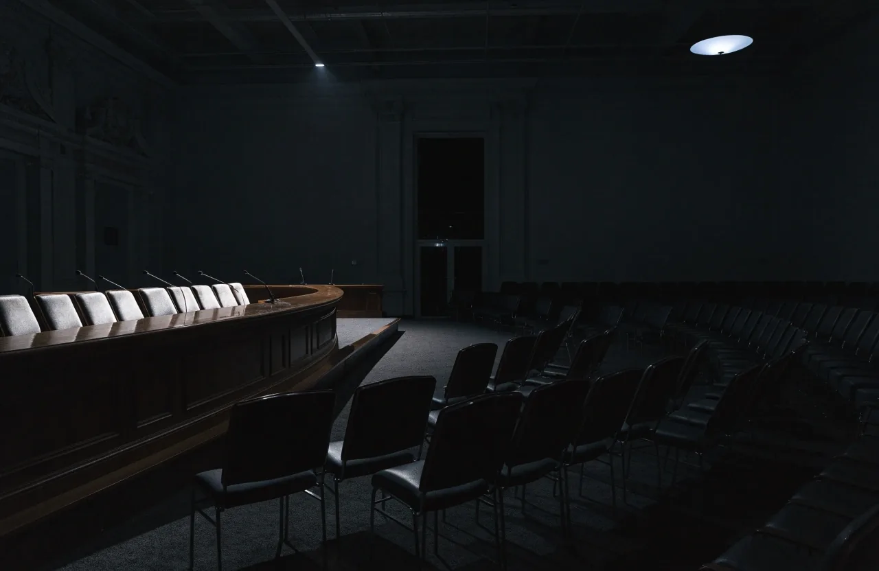 Empty hearing-room dais at night under a single overhead light, with vacant seating rows falling into shadow